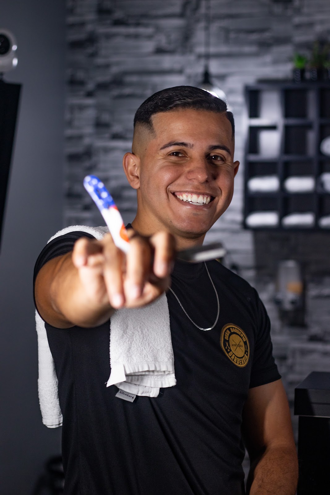 Barbeiro sorrindo, com toalha no ombro e o logo da barbearia na camiseta